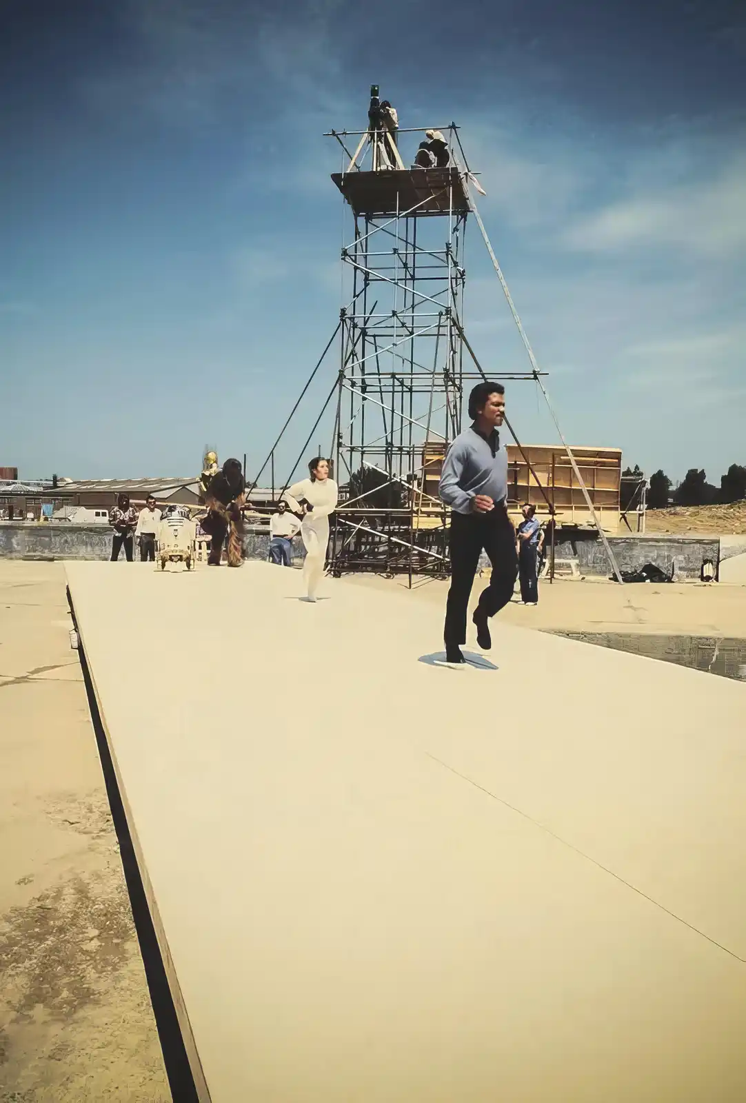 A behind-the-scenes photo from the Star Wars film set shows actors running on a large, flat, beige platform. In the…