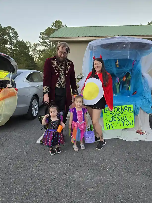 A family in Halloween costumes poses in a parking lot; a man as a pirate, a woman as a devil holding a fried egg, and two…
