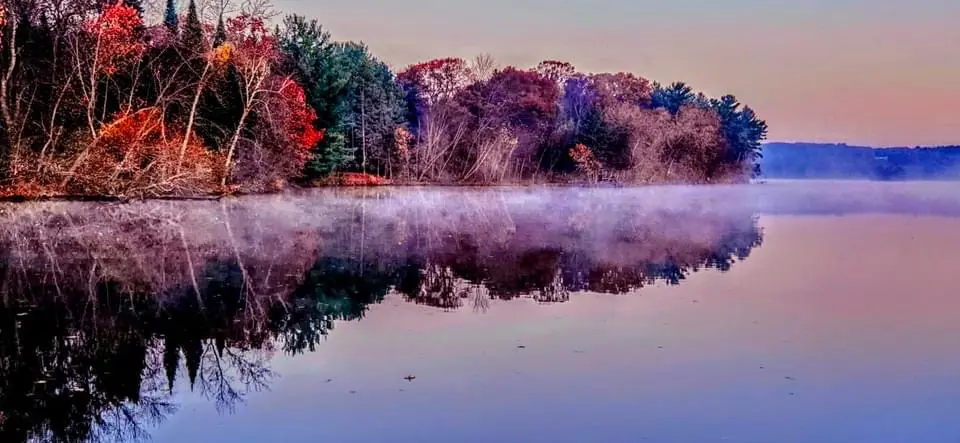 A serene, misty lake at dawn or dusk with colorful autumn trees on the shore, their reflections mirrored perfectly in the…