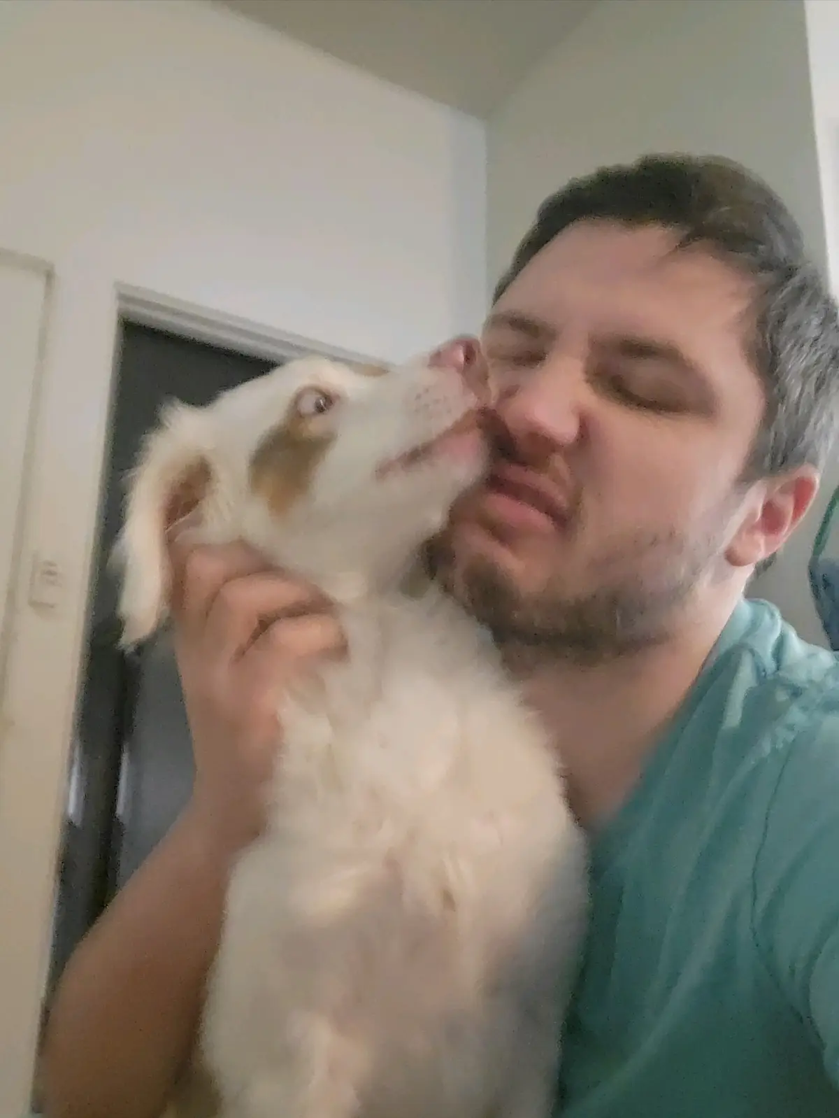 A man in a teal shirt holds a white and tan dog close, the dog licking his face while he closes his eyes with a blissful…