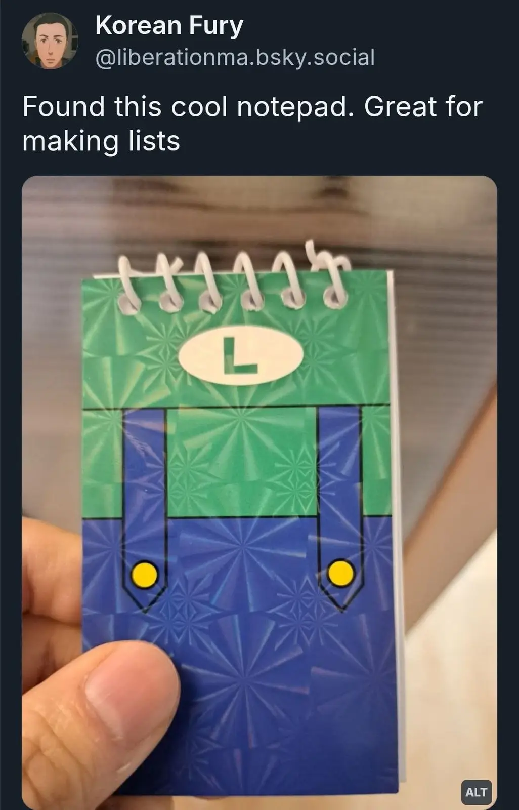 A hand holds a small spiral-bound notepad designed to look like the character Luigi from Mario games, featuring his green…