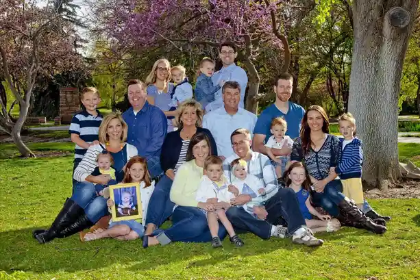 A large family poses outdoors under blooming trees for a portrait. A young girl in the front row holds up a framed photo of…