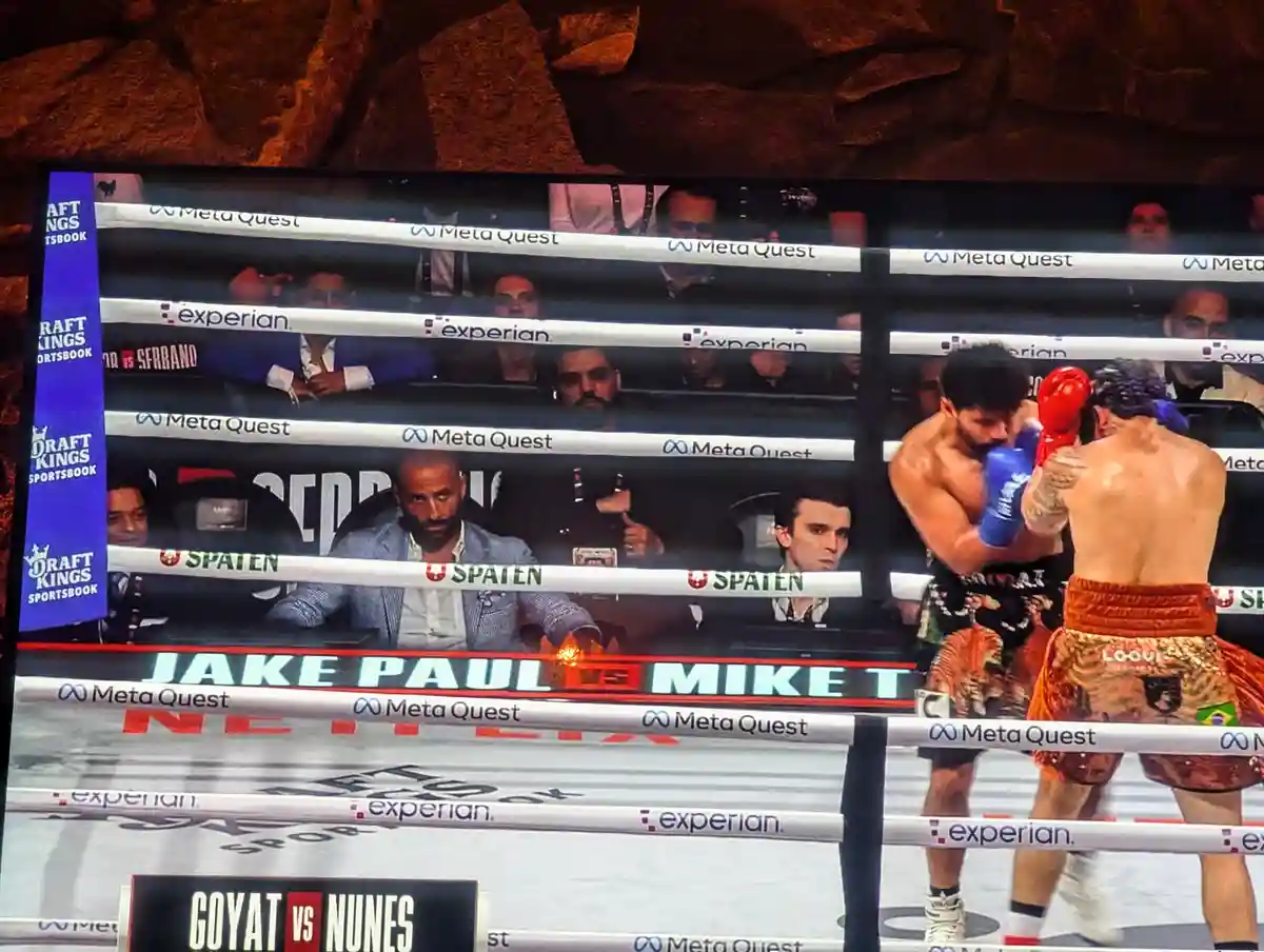 A television screen shows a boxing match between Jake Paul and Mike Tyson, with Tyson in red gloves and Paul in blue…