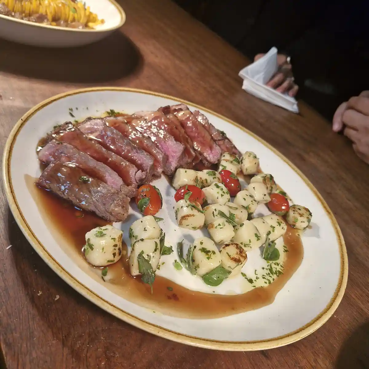 A plate of sliced medium-rare steak with gnocchi and cherry tomatoes, garnished with herbs and sauce, sits on a wooden…