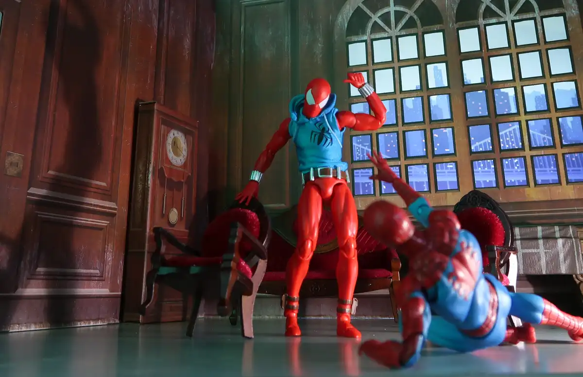 Two Spider-Man action figures are staged in a room with wooden paneling and large windows showing a cityscape. One figure…