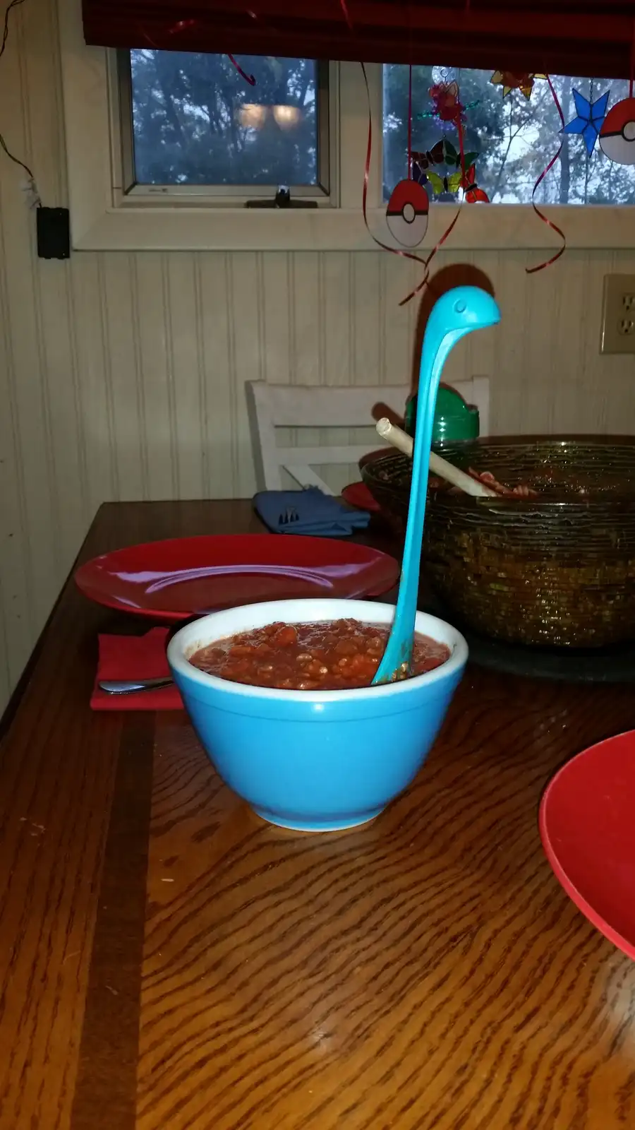 A blue bowl of red bean soup sits on a wooden table with a long-necked blue plastic spoon sticking out, resembling a…