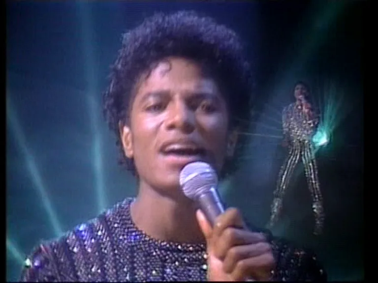 A close-up of Michael Jackson singing into a microphone, wearing a sparkly outfit, with a fainter, ghostly image of himself…