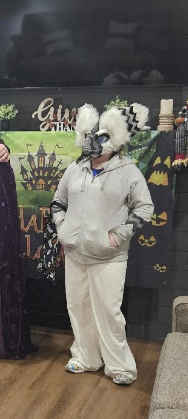 A person stands in a Halloween setting wearing a large, elaborate animal mask with white fur and black markings, paired with…