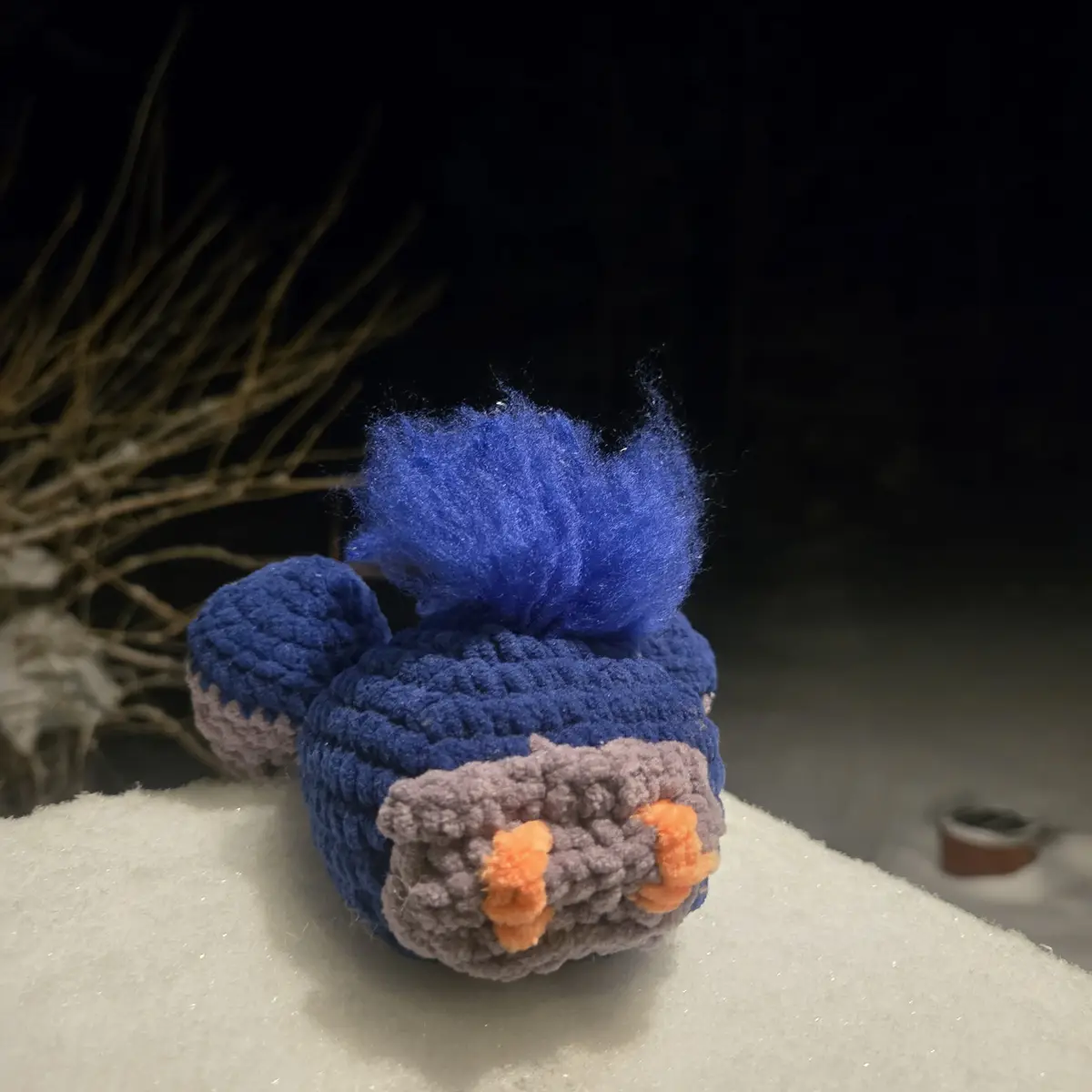A handmade crocheted plush toy resembling a blue bird with spiky blue hair, grey beak, and orange feet sits on a snowy…