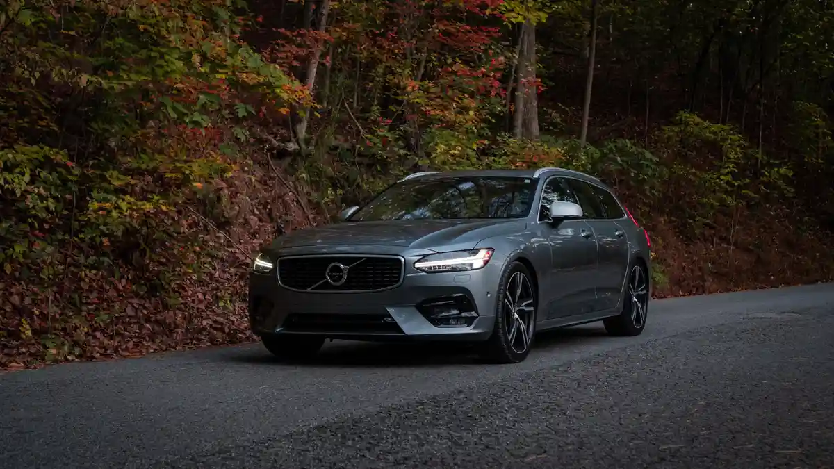 A gray Volvo station wagon is parked on a paved road next to a forest with autumn foliage. The car's headlights are on, and…