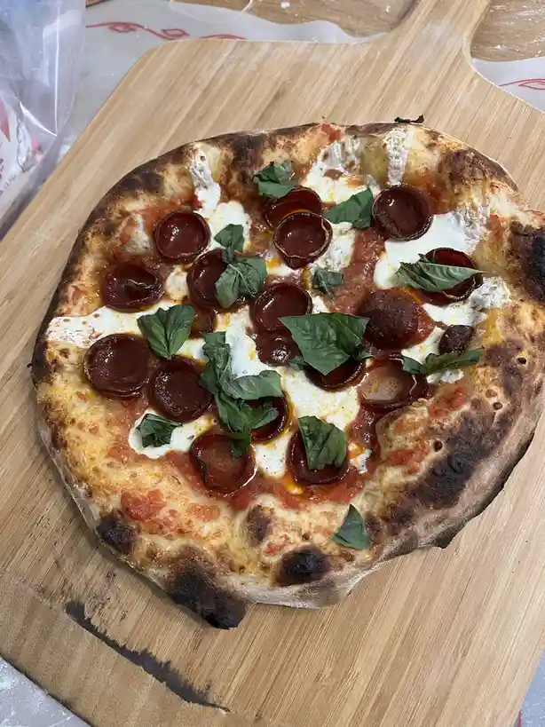 A close-up photo shows a round, rustic pizza with a charred crust, tomato sauce, melted cheese, pepperoni slices, and fresh…