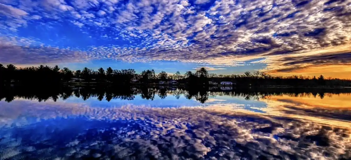 A serene landscape photo shows a calm lake reflecting a vibrant sky with blue and orange clouds at sunset or sunrise…