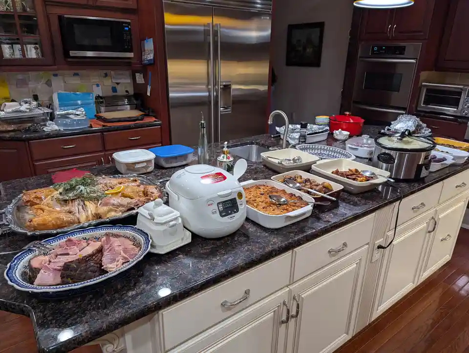 A kitchen counter is covered with a large spread of holiday food, including a roasted turkey, sliced ham, stuffing, roasted…