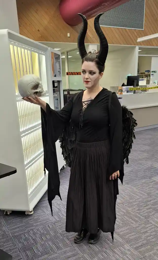 A woman dressed as a demon with black horns and feathered wings stands in an office or library, holding a human skull in…