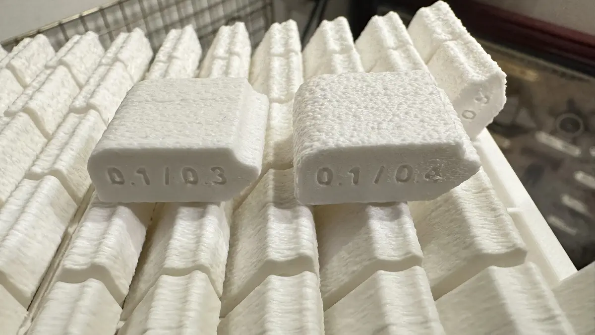 A close-up photo shows two white, textured, rectangular objects resembling 3D-printed plastic parts, each with an engraved…