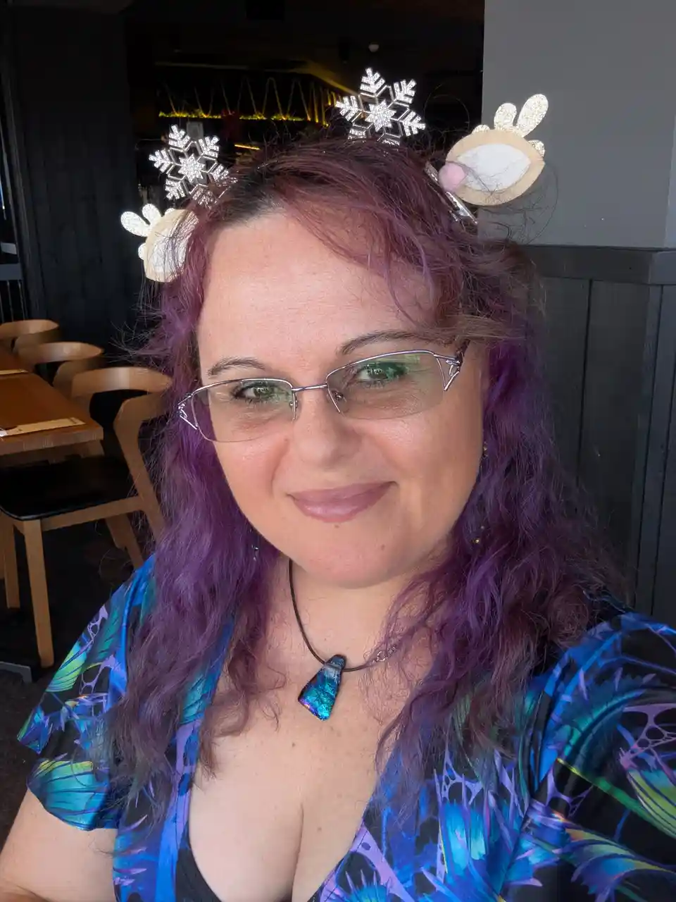 A woman with purple hair and glasses wears a festive headband with glittery snowflake and reindeer antler decorations while…