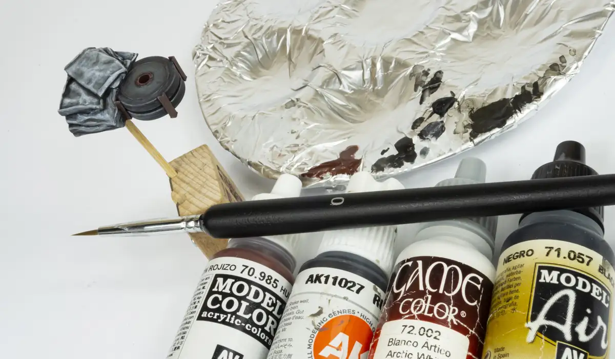 A still life of model painting supplies including acrylic paint bottles, a fine-tipped brush, a foil palette with black and…