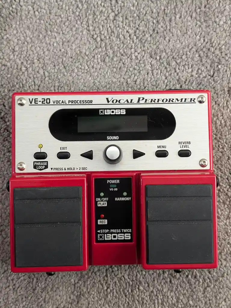 A red and silver Boss VE-20 Vocal Processor pedal sits on a gray carpet, showing its controls, display, and footswitches…