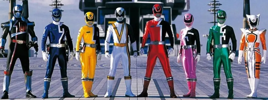 seven power rangers in full costume stand in a line, each wearing a distinct color suit with a number on their chest. The…