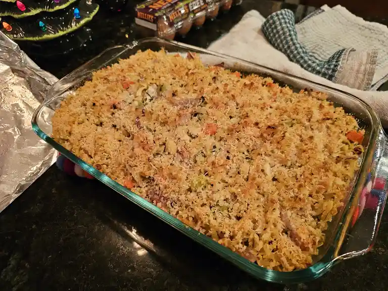A freshly baked pasta casserole with a golden-brown crumb topping sits in a clear glass baking dish on a dark countertop. In…