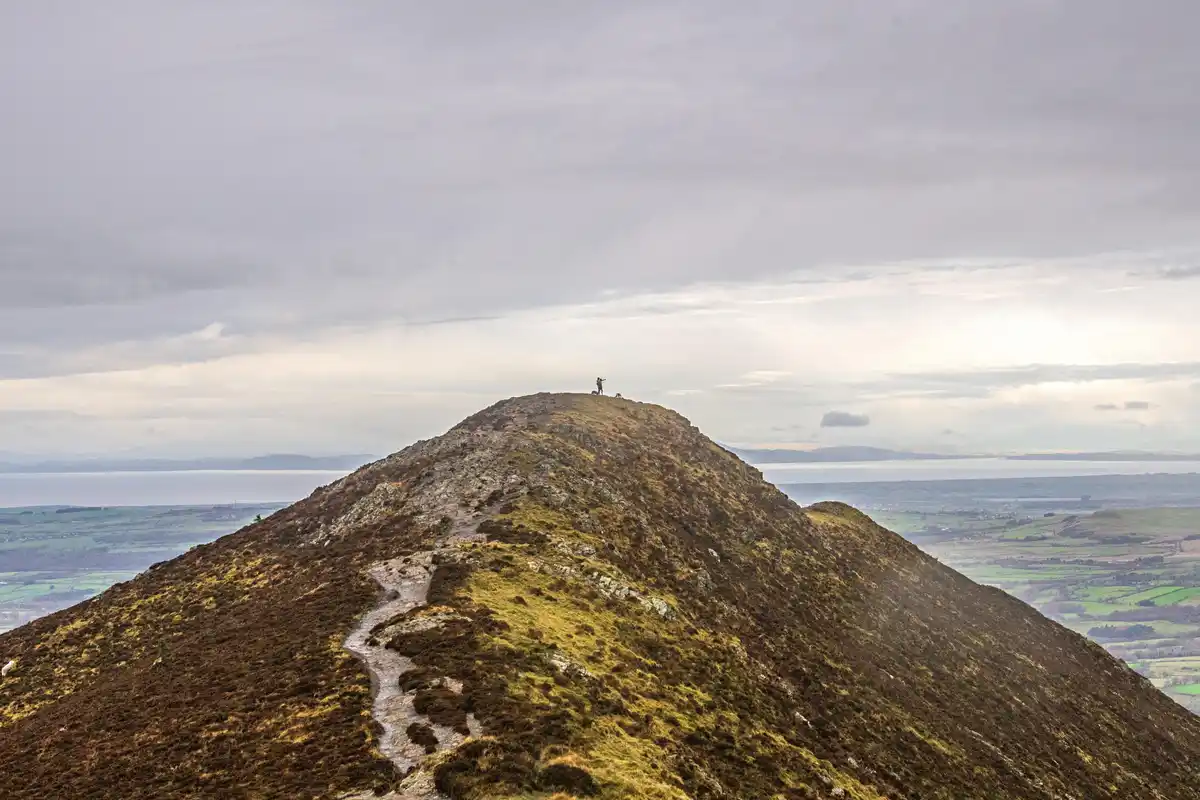 A lone hiker stands triumphantly atop a grassy, rocky hill under an overcast sky, with a vast landscape of fields and water…