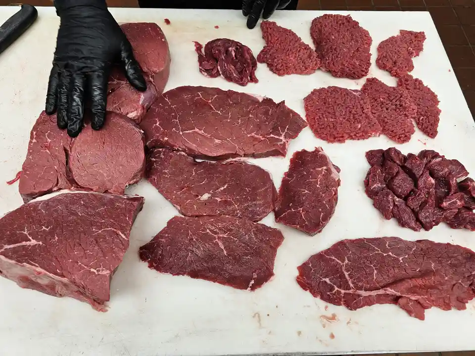 A person wearing black gloves arranges various cuts of raw red meat on a white cutting board, including large steaks, ground…