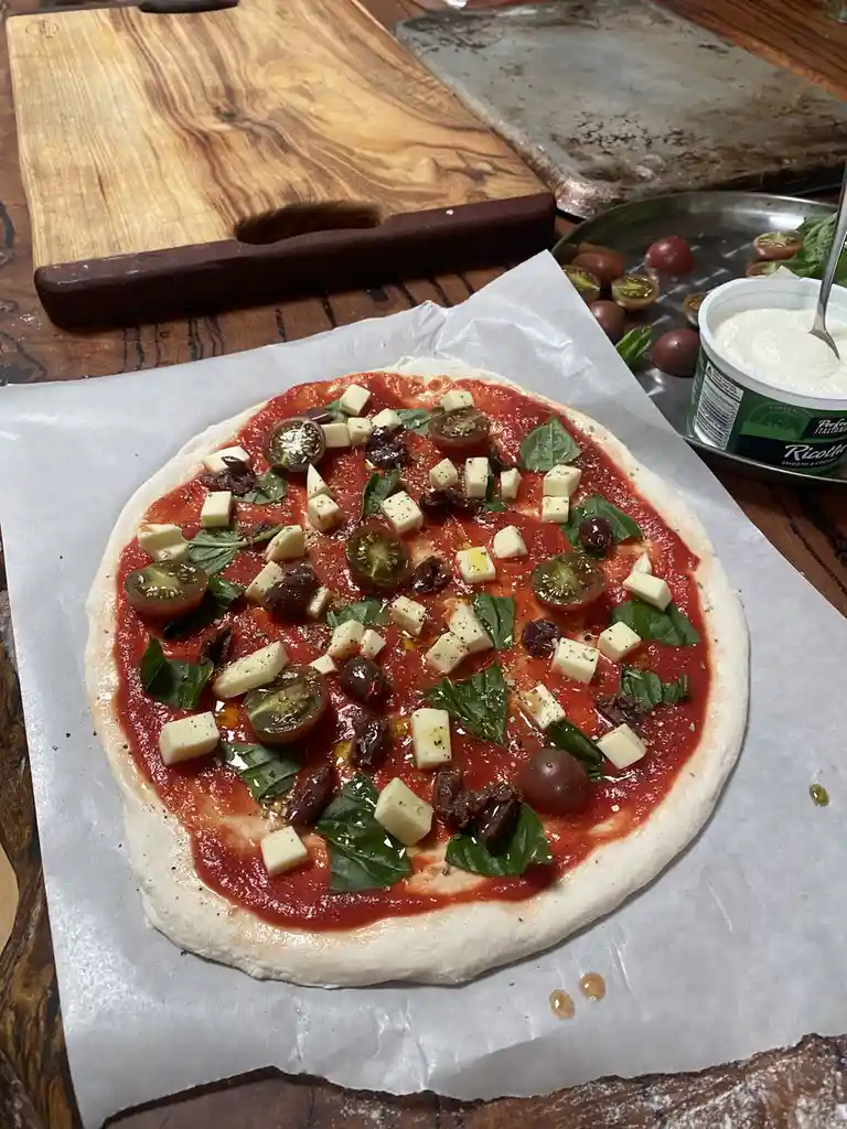 An uncooked pizza on parchment paper, topped with tomato sauce, cubed ricotta cheese, basil leaves, sliced cherry tomatoes…