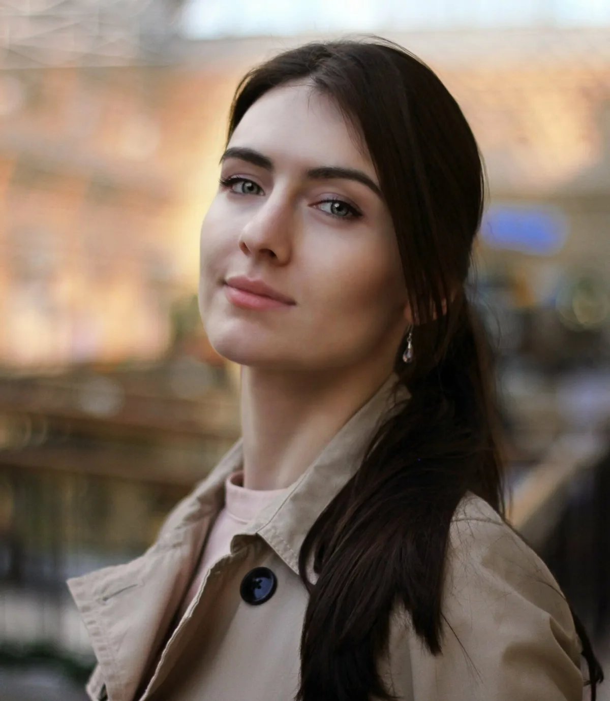 A woman with dark hair and green eyes, wearing a beige trench coat over a pink shirt, looks off to the side with a subtle…