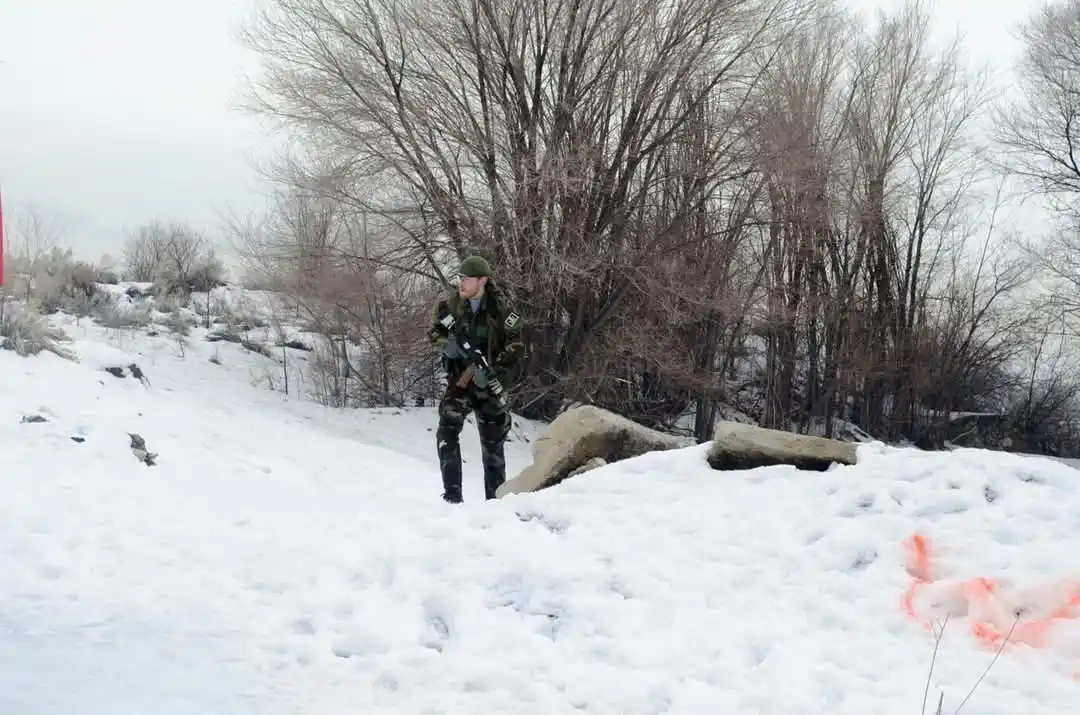 A person in full camouflage military gear, including a helmet and carrying a rifle, stands in a snowy outdoor environment…