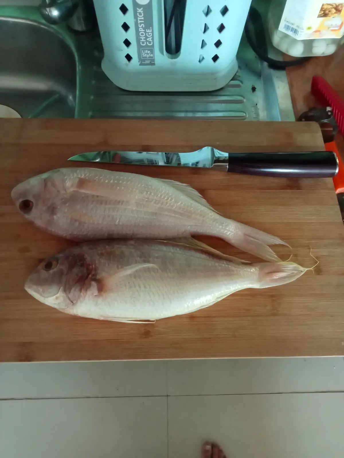 Two whole raw fish lie side-by-side on a wooden cutting board in a kitchen, with a knife resting above them. In the…
