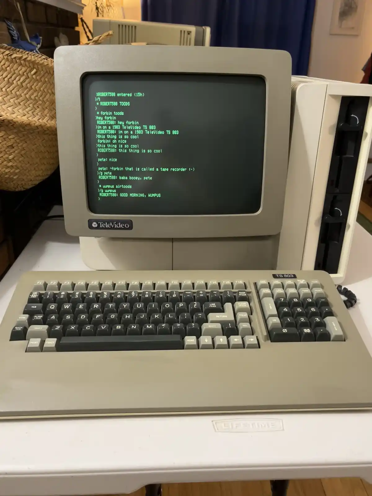 A vintage 1983 TeleVideo TS 803 computer with a beige monitor and keyboard, displaying a green text-based chat log on its…