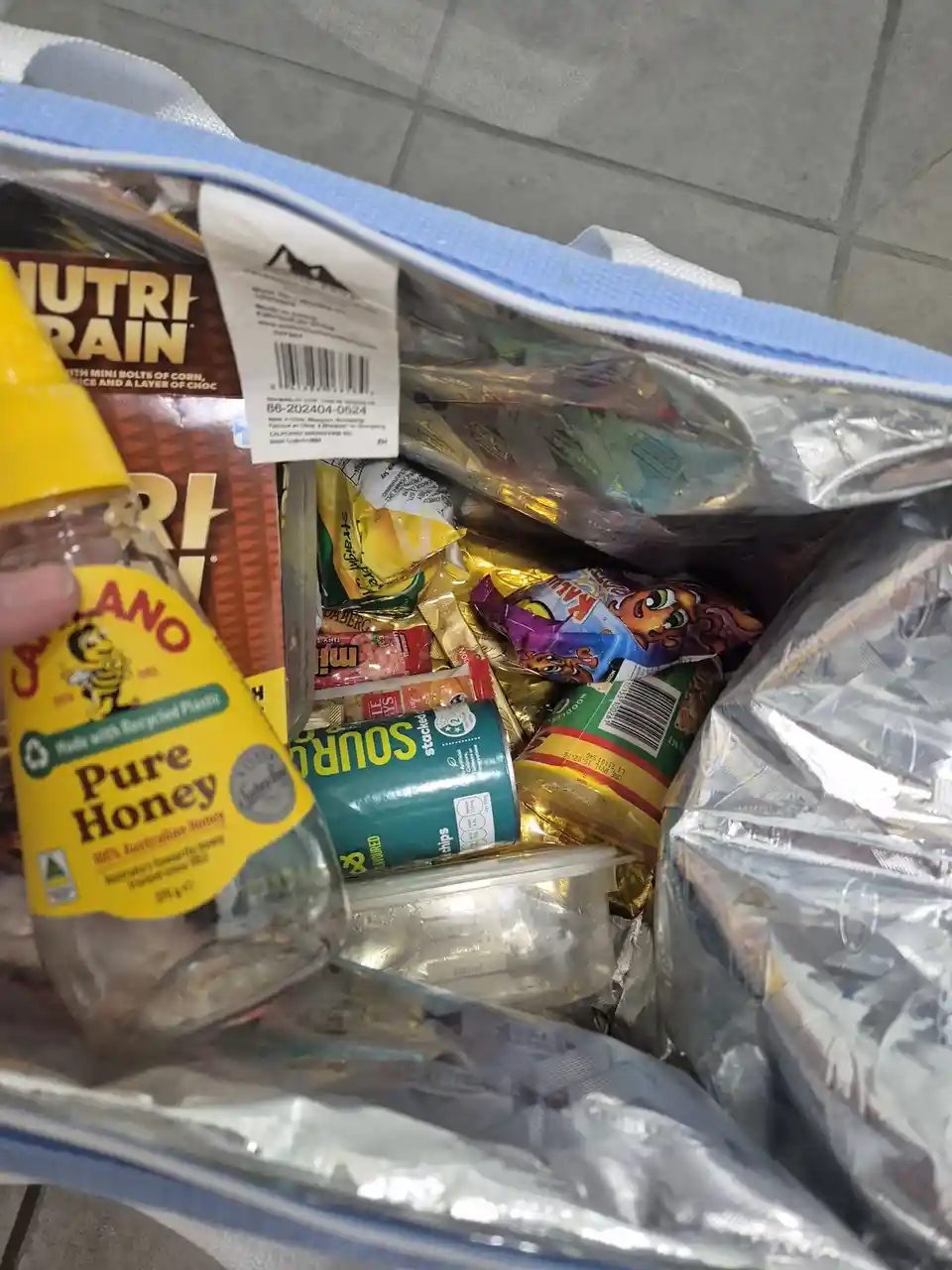 A close-up shot shows a person holding a bottle of Manuka honey in front of an open, silver-lined cooler bag overflowing…