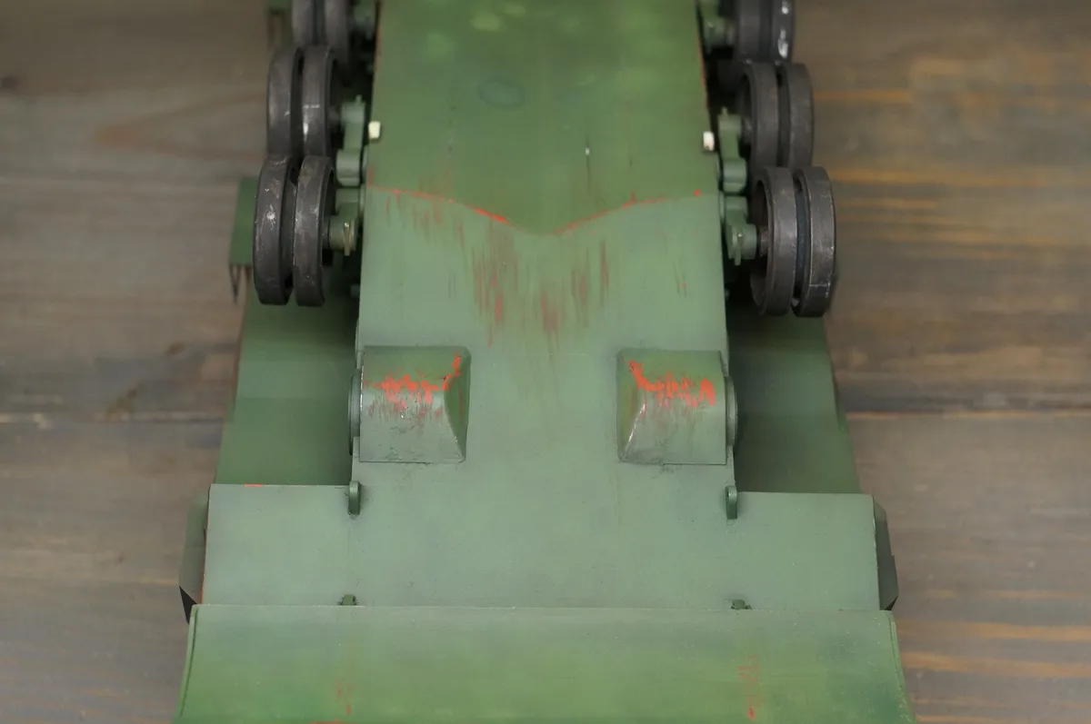 A close-up photo of the front of a green toy tank model on a wooden floor, showing its tracks, wheels, and a red paint drip…
