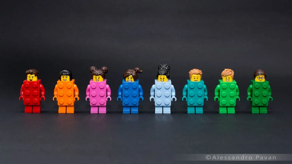 Seven Lego minifigures stand in a row against a dark background, each wearing a large Lego brick as a costume covering their…