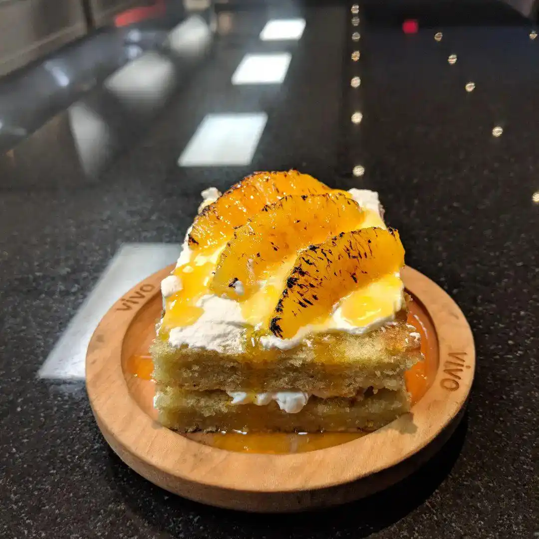 a slice of layered cake with white frosting and candied orange slices sits on a wooden plate branded with 'vivo', placed on…