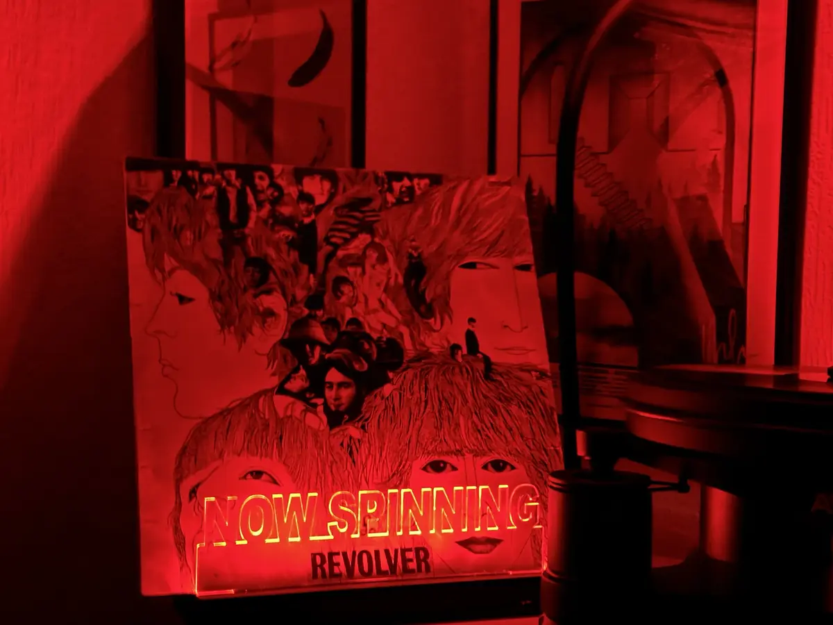 A red-lit vinyl record player with a custom album cover for The Beatles' 'Revolver' displayed prominently. The cover art…