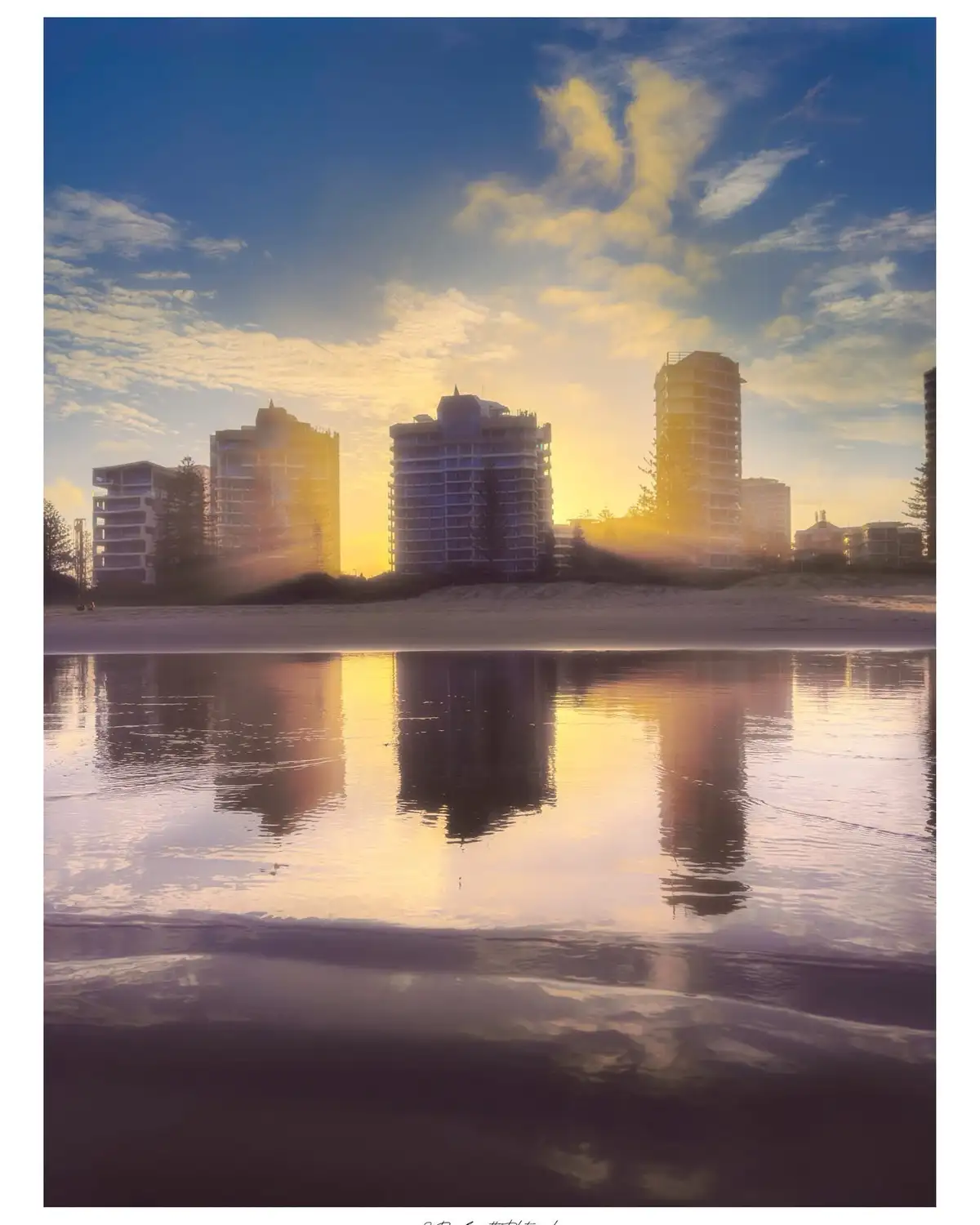A serene beach scene at sunrise or sunset, featuring tall coastal buildings silhouetted against a golden sky with scattered…