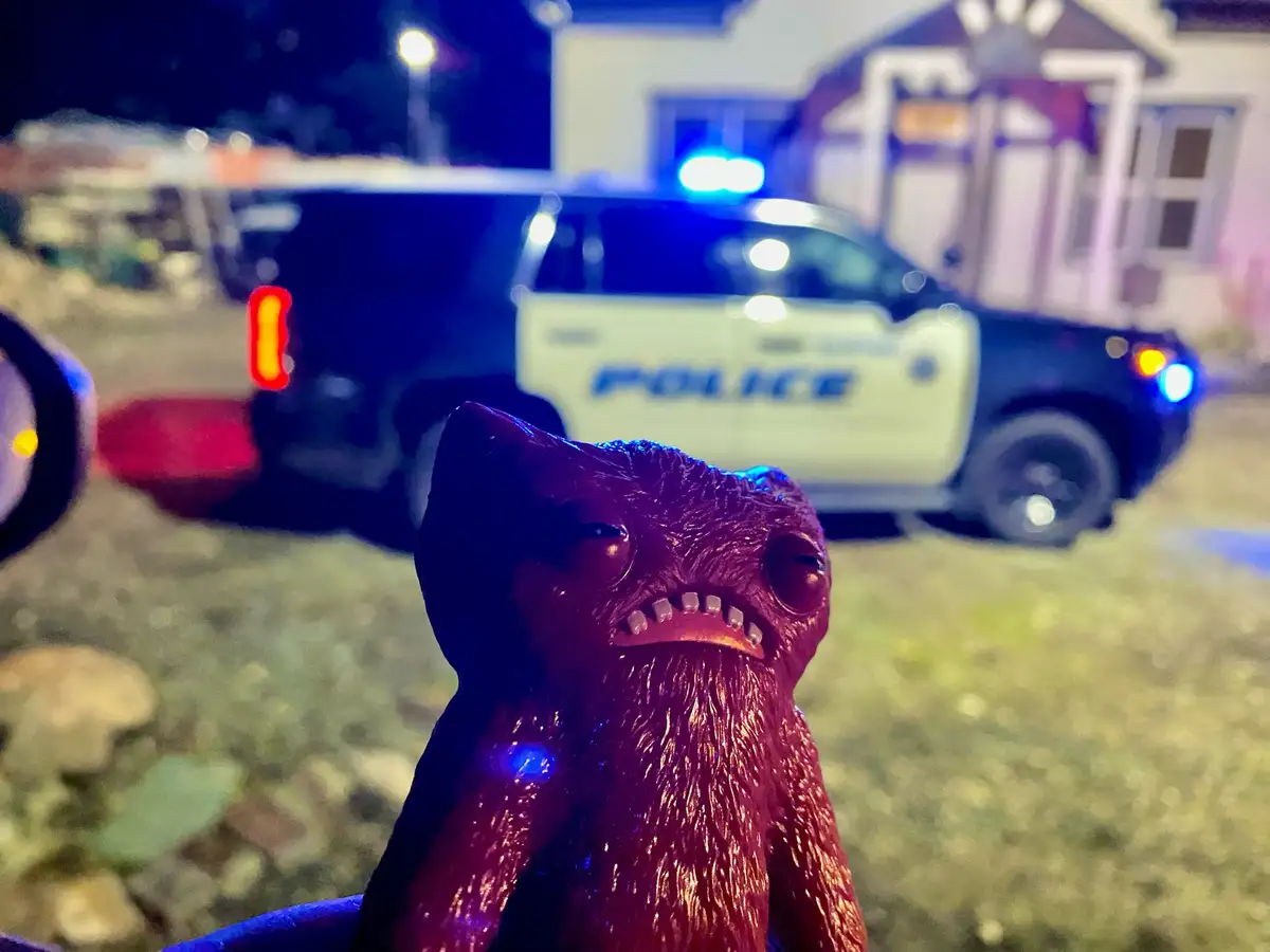 A close-up of a red, textured, cartoonish monster toy with a grumpy expression is in the foreground, with a blurred police…