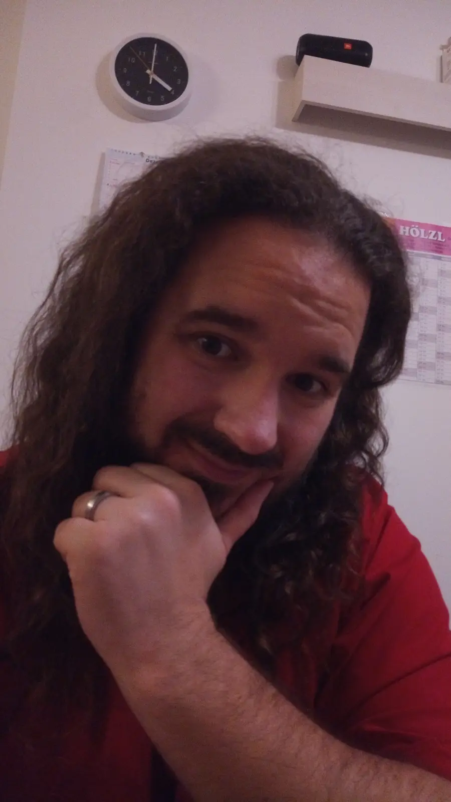 A man with long curly hair and a mustache, wearing a red shirt, rests his chin on his hand with a smug, thoughtful…