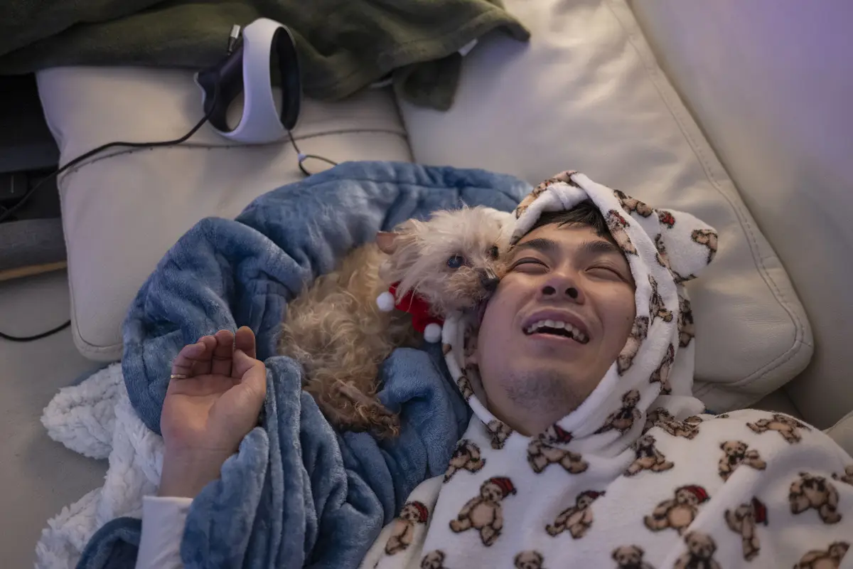 A person wearing a festive teddy bear-patterned robe with a hood is lying on a couch, smiling broadly with eyes closed while…