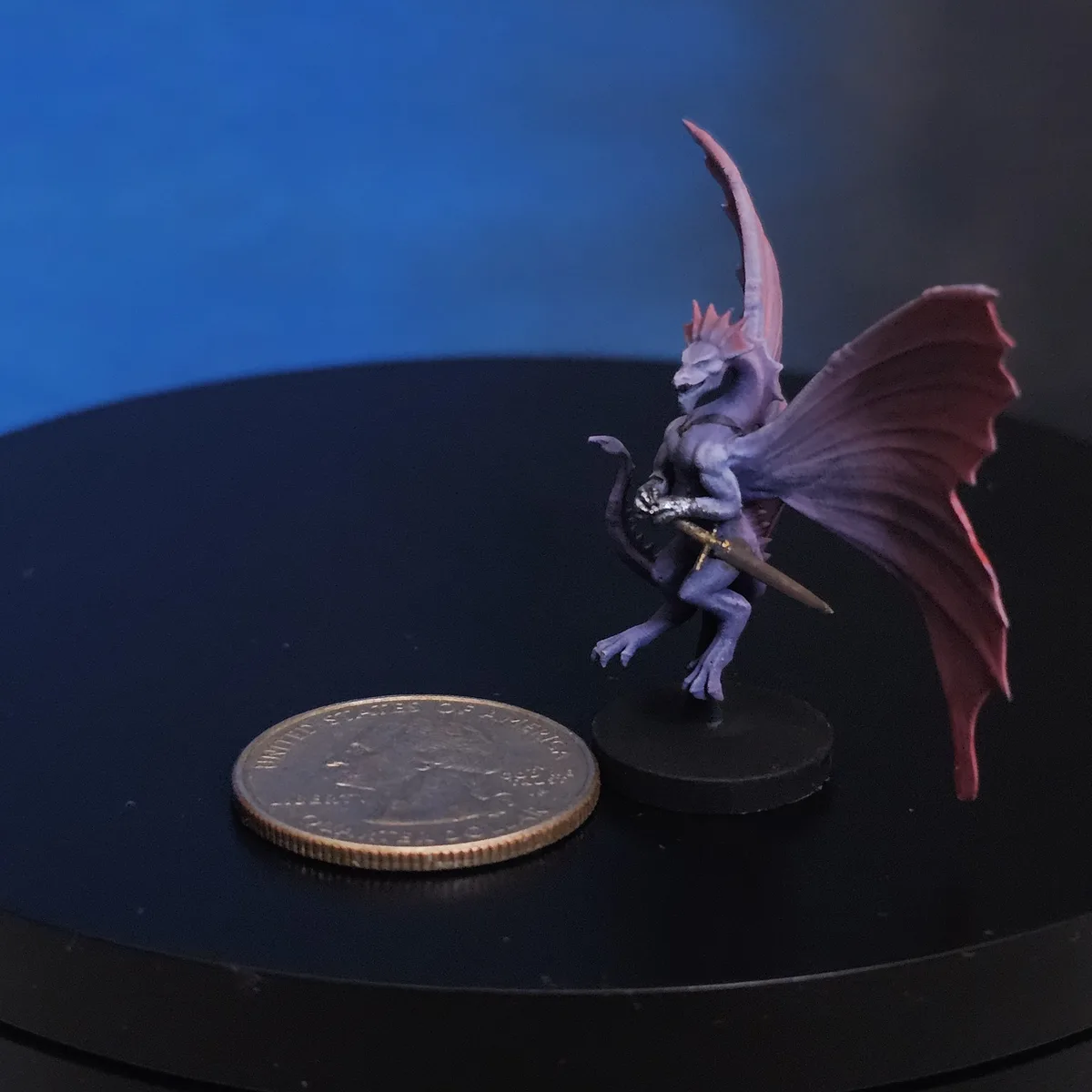A small, painted purple and red dragon miniature with wings and a sword stands on a black base next to a U.S. quarter for…