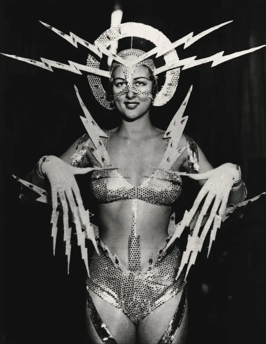 A black-and-white photo of a woman in a sparkly, perforated costume with a large, circular headpiece adorned with lightning…