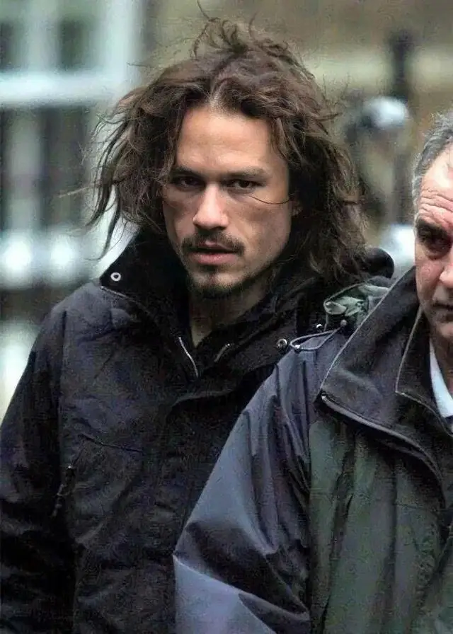 A man with long, messy brown hair and a goatee, wearing a dark jacket, stares intensely at the camera with a skeptical…