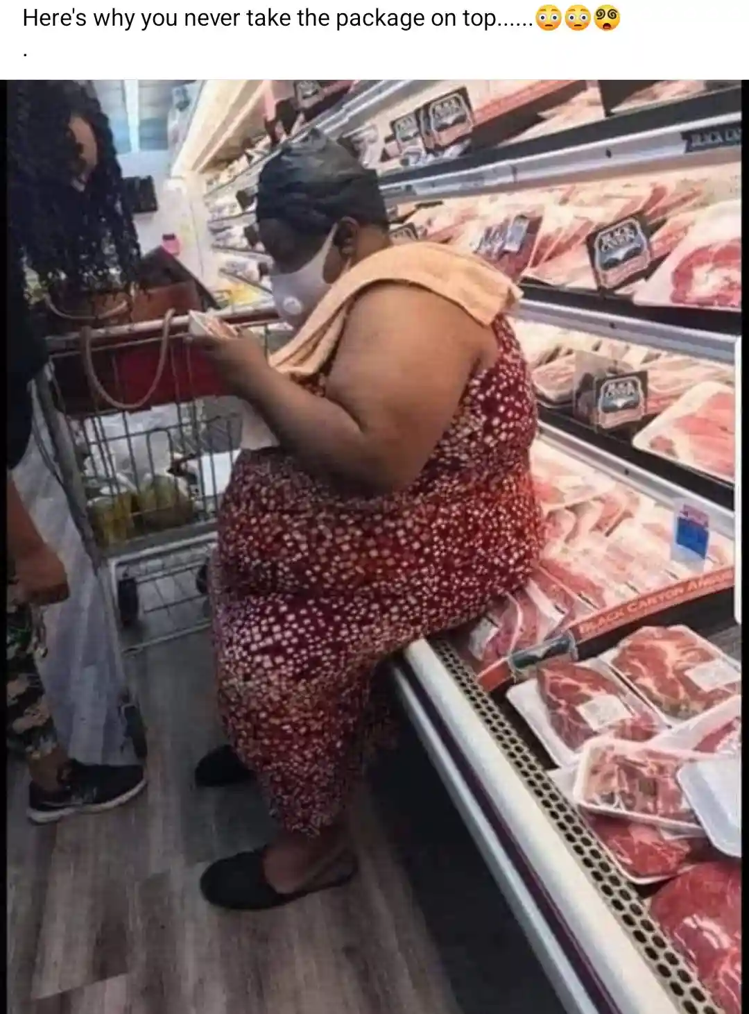 A person wearing a face mask and a red patterned outfit is sitting on a supermarket meat aisle shelf, leaning over a…