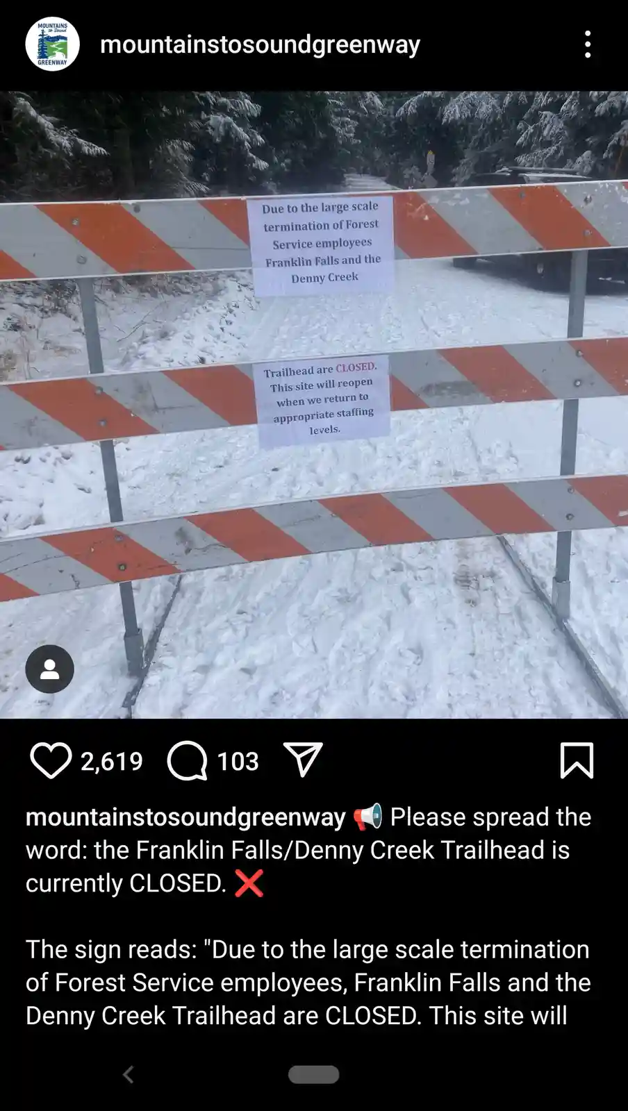 A snowy trailhead is blocked by orange and white striped barricades with two handwritten signs. The top sign states the…