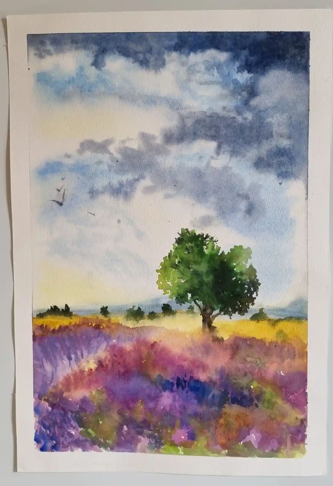 A watercolor painting depicts a lone green tree standing in a field of purple and pink flowers under a cloudy blue sky with…