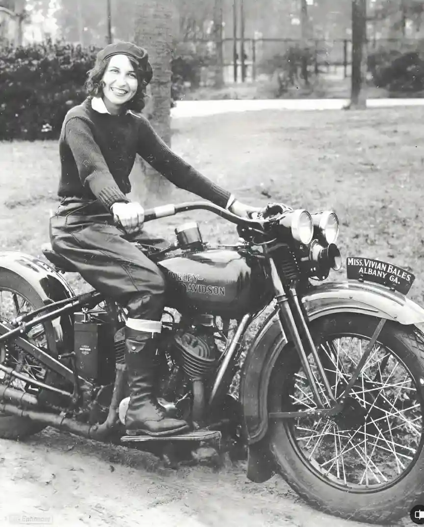 A vintage black-and-white photo shows a smiling woman in a beret and sweater riding a Harley-Davidson motorcycle. The front…