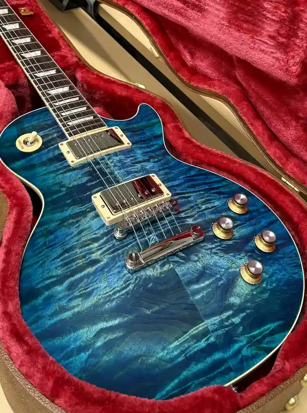A close-up photo of a blue electric guitar with a swirling, flame maple finish, resting inside a plush red-lined case. The…