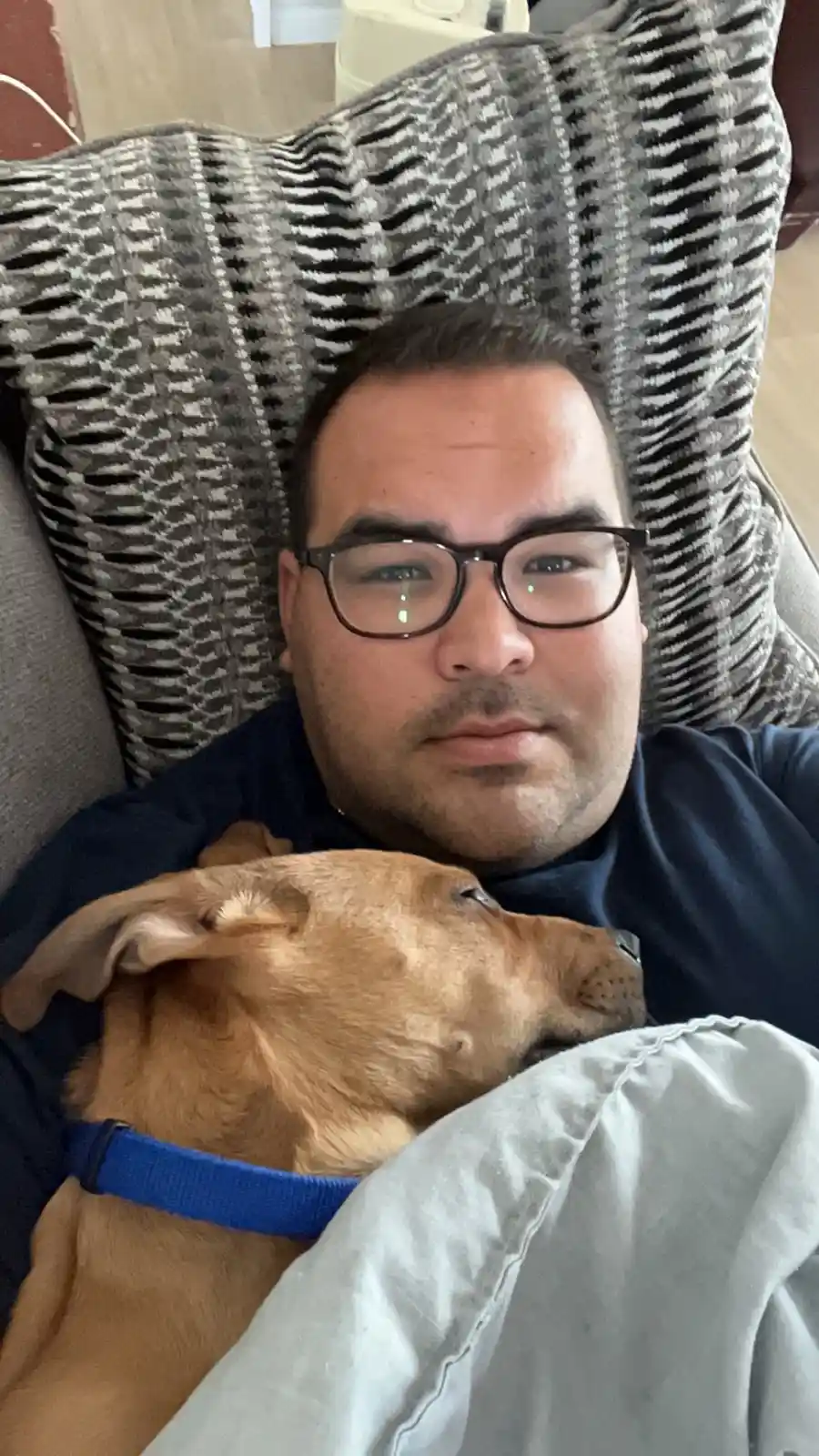 A man with glasses lies on a couch, looking directly at the camera with a neutral expression, while a tan dog with a blue…