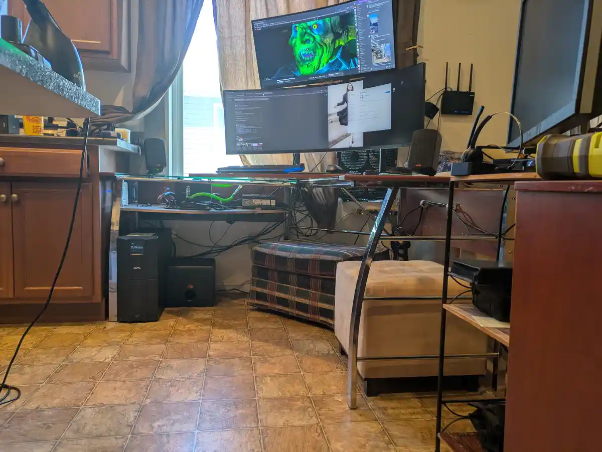 A cluttered home office setup with multiple monitors, one displaying a green-skinned horror movie character, surrounded by…